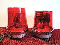 Pair of vintage emergency lights side by side on red textured surface, showing red plastic domes and metal bases with labels and wiring.