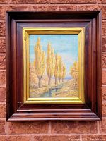 Full view of the framed landscape oil painting showing yellow autumn trees by a stream under a blue sky, set against brick wall.