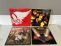 Four vinyl albums arranged in pairs, top left Aerosmith Greatest Hits in red and white cover, top right Genesis album with cube art, bottom left Nazareth with picnic cover, bottom right Triumph Eliminator album