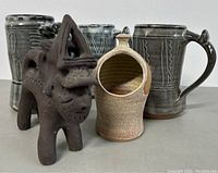 Three handmade ceramic mugs with different decorative patterns and a small dark brown South American style animal figurine, plus a pottery container with a spout and handle.