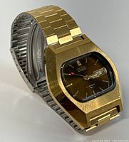 Side angle showing gold-tone case and two-tone bracelet with brushed finish, displaying the watch face with dark brown dial, date/day window, and red second hand.