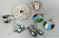Lot of sterling silver jewelry showing earrings, ring, and brooch together on gray background. Includes various stones and abalone shell inlay earrings, peach stone ring, and engraved brooch.