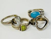 Four small sterling silver rings. Three have stones (clear round, rectangular green, oval turquoise), one open heart shaped band. Visible tarnish on all.