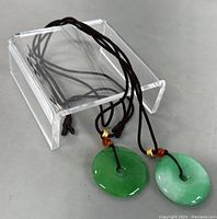 Two jade disk pendants and braided cords displayed on clear acrylic stand.