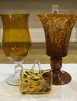 Photo showing tall amber blown glass pedestal vase next to shorter tortoise shell pedestal vase and amber tortoise shell blown glass purse figurine on white surface.