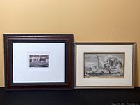 Two framed prints side by side against a beige wall, one dark wood frame with a small landscape print, one lighter frame with a classical ruins print.