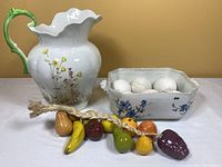 Floral porcelain pitcher with green handle next to rectangular porcelain dish containing white eggs and porcelain fruit garland on white surface.