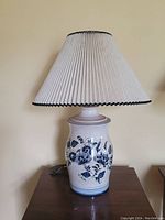 Full view of a porcelain decorative table lamp with blue floral design and pleated white fabric shade with black trim.