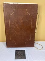 Brown leather slipcase with gold rope tie beside square metal bas-relief sculpture showing figure motif