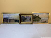 Three framed landscape paintings on board arranged on a table, showing lakes, boats, trees, and mountains.