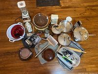 Lot overview showing all items laid on wooden floor: crockpot, blender, coffee grinder, cake mold, bean pot, pans, baking tray, electric can opener, utensils.