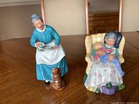 Both Royal Doulton figurines displayed on a wooden table. One figurine is an elderly woman with a cat, the other is an elderly woman with a child seated in an armchair.
