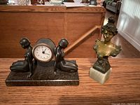 Front view of Deco sculpture clock showing two kneeling nude figures in bronze on dark marble base next to Art Nouveau bronze bust on light marble base