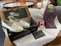 Display of all items: white and beige Callaway Rhiona golf shoes new in box, stainless steel Mapleleaf birdie juice flask with maple leaf, purple Goose Golf towel, and putting mate set on white cloth.