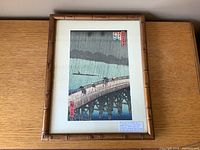 Framed Japanese woodblock print with bamboo-style frame showing rain and bridge scene.