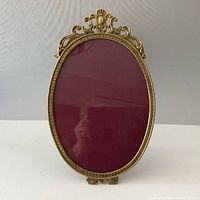 Front view showing ornate brass oval frame with decorative top and red backing behind glass.