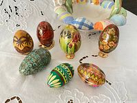 Multiple painted Easter eggs displayed on white embroidered cloth with a colorful ceramic basket in background. Various designs and mediums including brass stands visible.