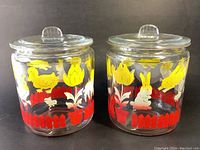 Two glass canisters with lids decorated with yellow and white animals, red picket fences, and flowers, side by side against a black background.
