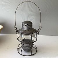 Front view of vintage metal railroad lantern showing clear glass globe, wire protective cage, and wire handle.