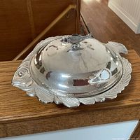 Photo showing full covered dish with fish-shaped handle on the lid, reflective silver-plated surface, and fish design details on the base.