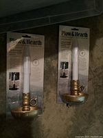 Two packaged Plow & Hearth battery operated candles in brass holders with finger ring handles visible in clear plastic and branded cardboard backing.