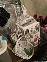Photo showing white decorative bird cage with ornate roof, painted wooden birdhouse with floral design, and white wicker basket in front.