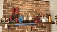 Decorative items on a wooden shelf against a brick wall including birdhouse, candleholders, pinecones, metal pitcher, and more.