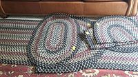 Six oval braided rugs piled on a larger patterned carpet, showing colors black, gray, cream, burgundy, and dark blue.