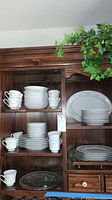 Wood cabinet with multiple shelves holding the china dishes, cups, saucers, plates, bowls and glass serving dish visible.