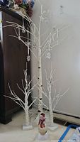 Three white birch-style lighted trees with hanging snowflake ornaments on bases, medium and large sizes visible