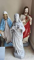 Three religious statues in a corner; Sacred Heart Jesus, Mary statues show chips and wear