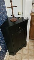 Black wooden corner cabinet placed in corner near door, showing one drawer and cabinet door with knob handles and visible surface scratches.