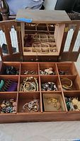 Open wooden jewelry box showing multiple compartments and mirrored doors with assorted costume jewelry inside including bracelets, necklaces, and pins.