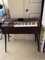 Full view of the vintage Magnus electric chord organ standing against a wall, showing wooden cabinet, 37 keys, and chord buttons on top and side.