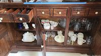 Wooden cabinet showing various glassware, ceramic items including soup tureen, mugs, punchbowl, glasses, serving platter, candlestick with flameless candle