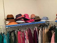 Shelf with seven ladies hats of various colors and styles above hanging women's clothing.
