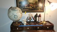 Wide shot of vintage globe, Lenox vase and pitcher, Hartmann's carved figures, gooseneck lamp, and Christmas decorations arranged on brown wooden stand below horse painting.