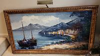 Framed signed coastal landscape painting showing boats on the water with a village and mountains in background