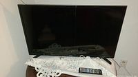 Front view of Samsung 32 inch TV sitting on a table with a white lace cloth, remote control placed in front.