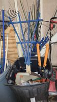 Lot of garden and yard tools, including blue wooden trellis, various hand tools with black and colored handles, seed spreader, and rakes leaning in the background.
