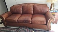 Full front view of brown leather sofa with three seat and three back cushions, metal rivet trim, placed in living room on a rug.