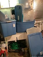 Photo of assorted plastic storage bins and totes stacked in basement showing variety of sizes and colors including blue and translucent bins.