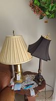 Two table lamps on a wooden side table. One lamp has a brass-colored metal base with a beige pleated shade, the other has a dark ornate metal base with a black lampshade.