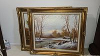 Two ornate gold-toned framed landscape prints leaning against a wall. Scenes depict wintertime or early spring with barren trees, water, and rocks. Both prints signed.