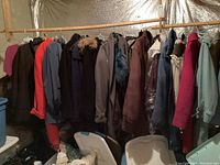 Photo of multiple women's winter coats hanging on a rod in a basement area. Various colors including dark blue, red, purple, brown, and pink. Some coats have fur trims on hoods.