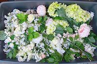 Top view of mixed silk flowers inside a plastic storage bin showing white, pale pink, and light green flowers with green leaves.