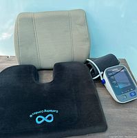 Photo of all items: Omron blood pressure monitor with cuff, beige back cushion, black seat cushion with blue embroidery