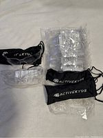 Nine individually wrapped clear plastic kids safety glasses arranged in a stack with three black soft drawstring cases branded 'ACTIVEKYDS'.