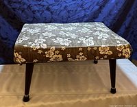 Rectangular Mid-Century Modern ottoman with Hawaiian floral fabric cover, showing wooden legs with metal foot caps and fabric condition under indoor lighting.