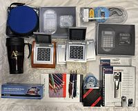 Wide shot showing various office accessories including desk clocks with calculators, digital cameras, pens, pencils, tape dispenser, travel mug, and USPS scale in packaging.